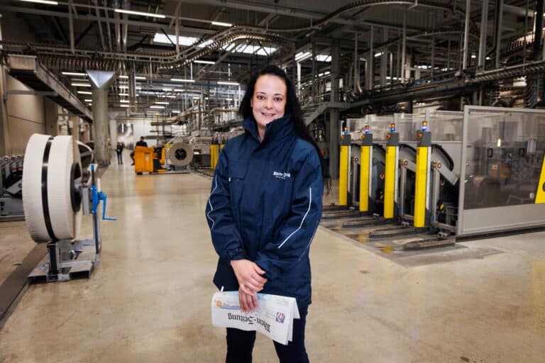 Eine Frau steht in einer Druckerei, lächelt in die Kamera und hält eine Zeitung in der Hand